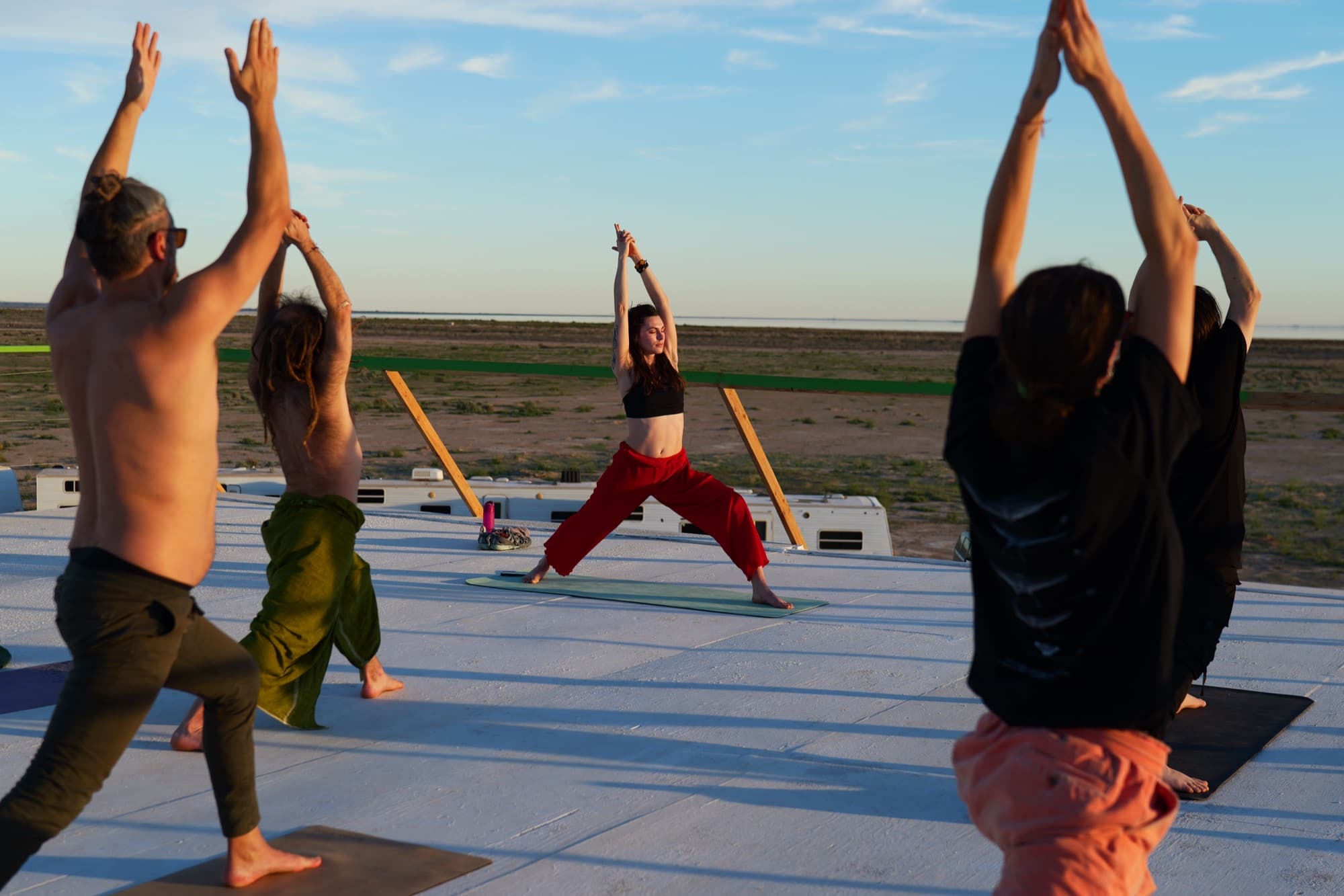 Julia leading a class in a wide warrior pose on a desert rooftop at sunset, students reaching up in the foreground